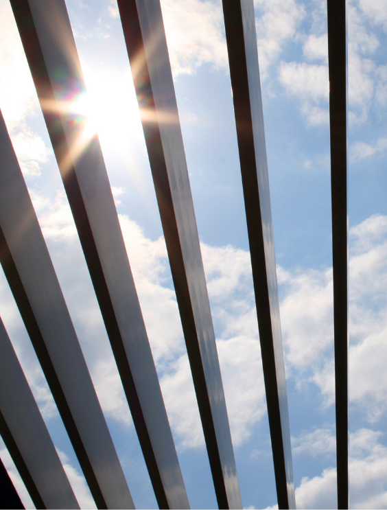 Een blauwe lucht met wolken en enkele verticale latten van en zwartgekleurde pergola waar de zon door schijnt