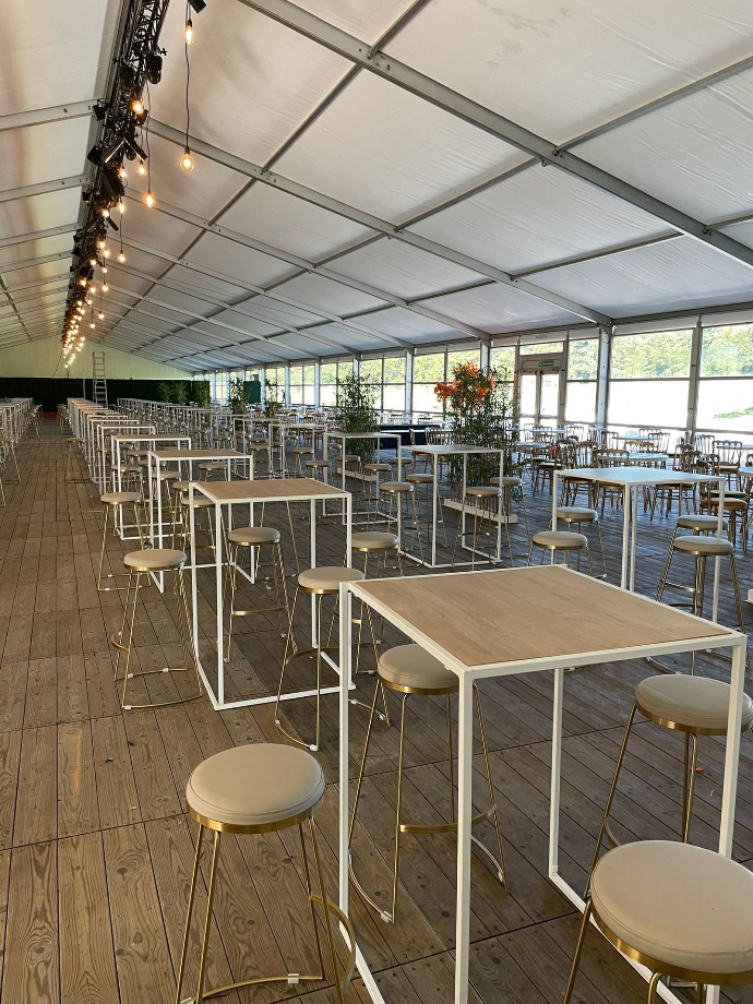 De binnenkant van een grote witte tent met houten vloer die gevuld is met hoge tafel en barstoelen met metalen, gouden details 